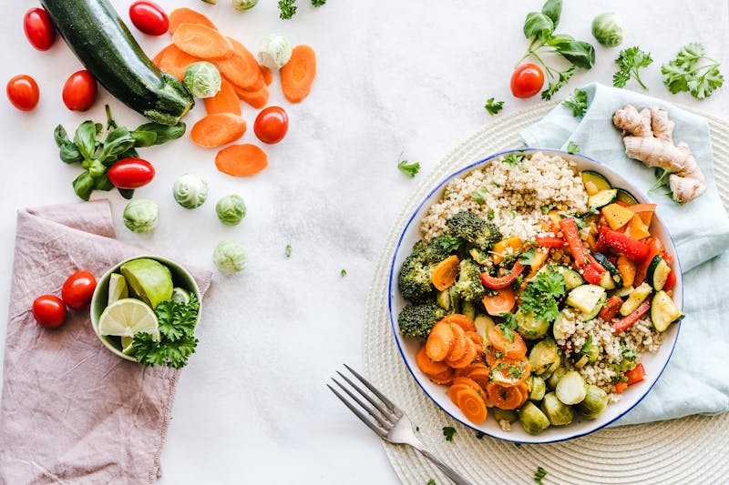 Bowl de verduras salteadas con quinoa junto a ingredientes crudos dispersos: tomates cherry, zanahoria, brócoli, limón y hierbas frescas sobre superficie blanca
