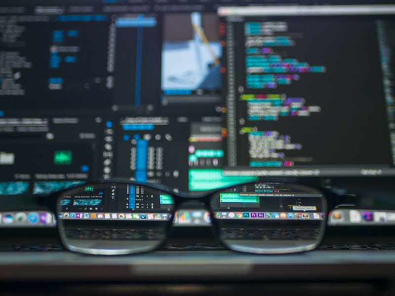 Gafas de montura negra en primer plano sobre una superficie, con código colorido desenfocado en la pantalla del fondo