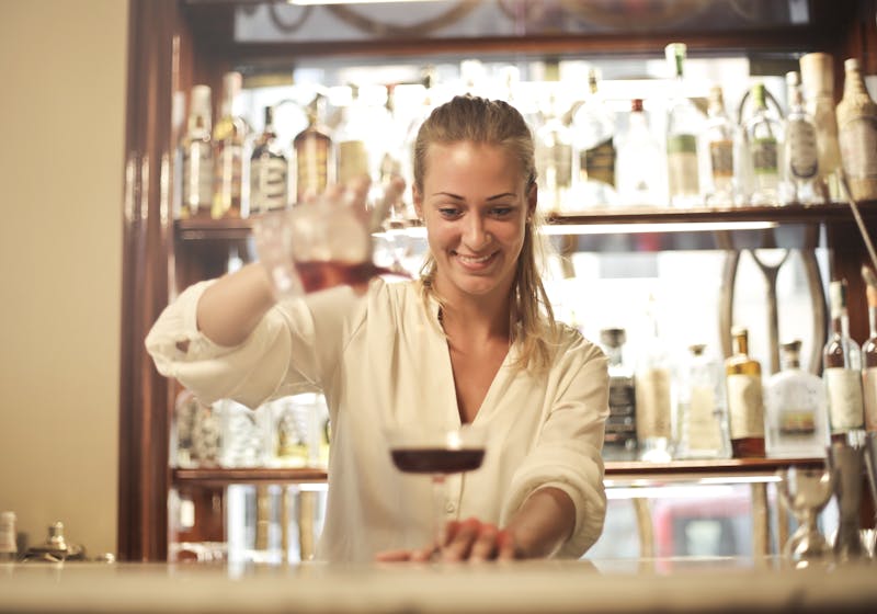 Bartender mujer de blanco sirviendo con precisión una copa de vino tinto en un bar elegante con estanterías de botellas al fondo
