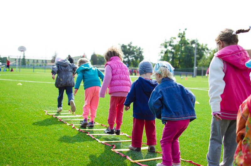 Grupo de niños en fila pisando uno a uno los peldaños de una escalera de agilidad sobre un campo de hierba