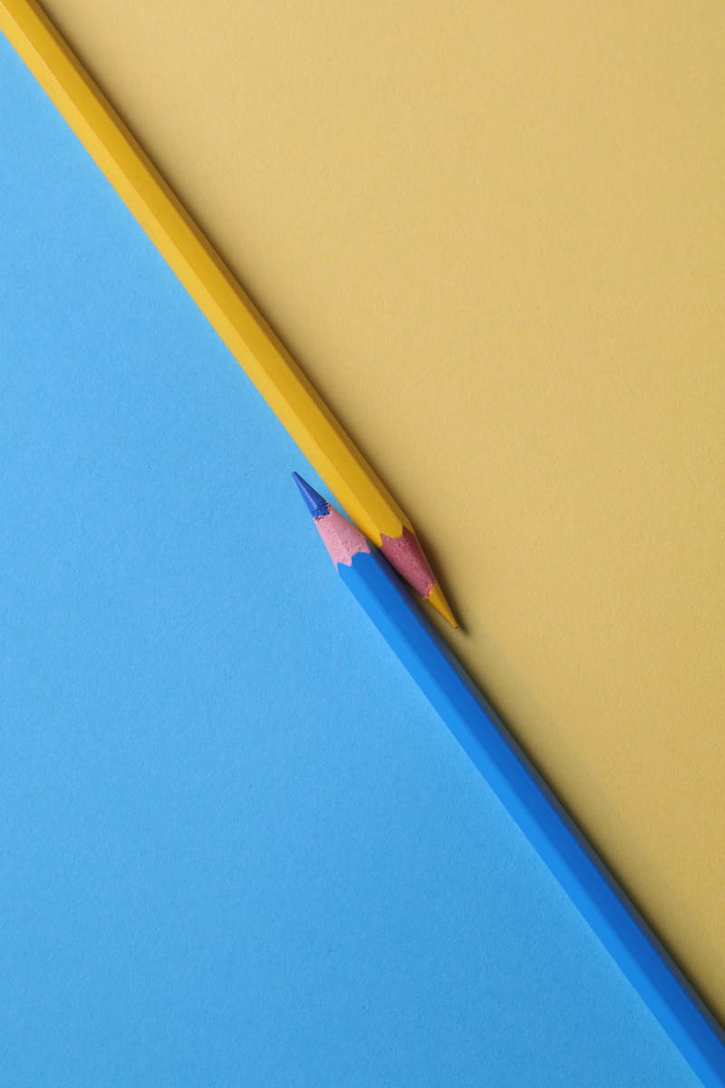 Un lápiz amarillo y un lápiz azul cruzándose en diagonal sobre un fondo dividido en los mismos dos colores