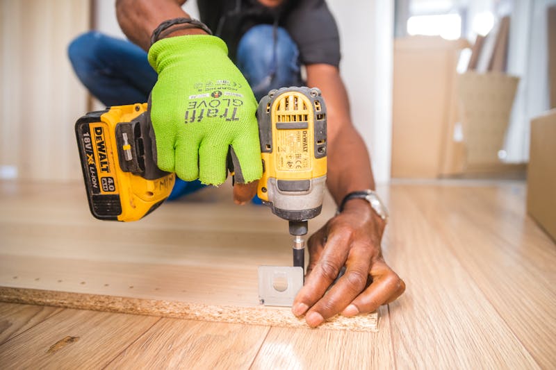 Mano con guante verde sosteniendo un taladro amarillo DeWalt sobre madera