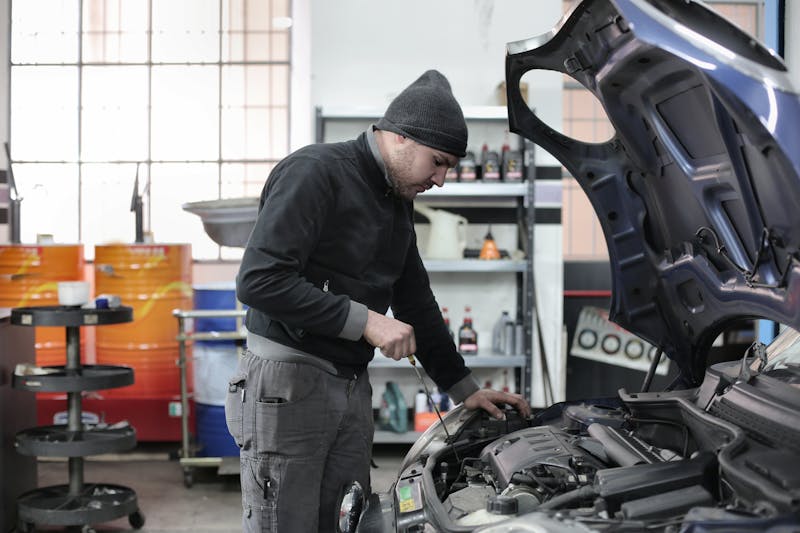 Mecánico trabajando bajo el capó de un coche en un taller con herramientas y estanterías al fondo