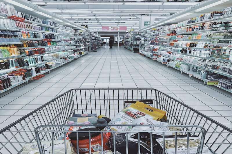 Pasillo de supermercado con estanterías llenas de productos y carrito de compra en primer plano