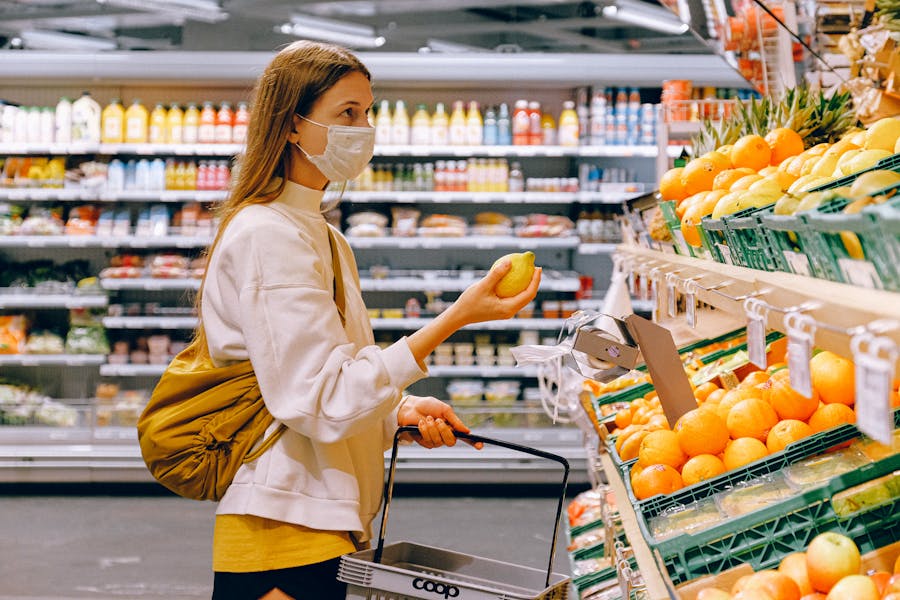 Mujer con mascarilla seleccionando productos de una estantería de supermercado con frutas y hortalizas