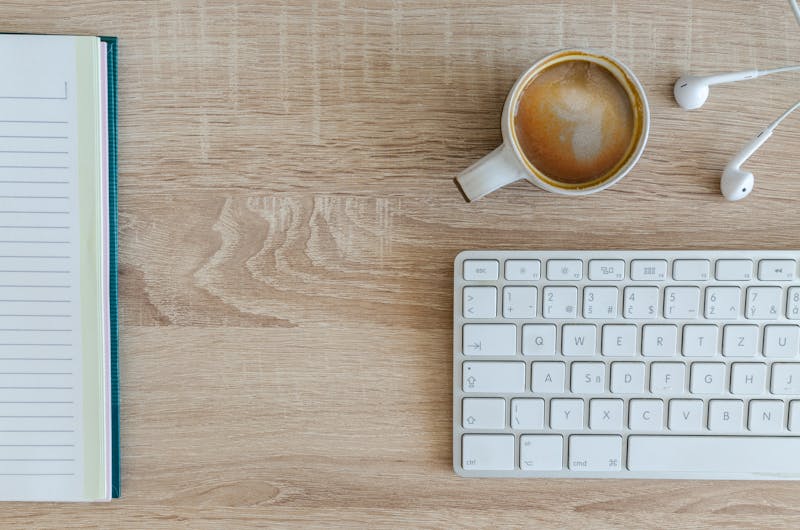 Libreta abierta junto a un teclado y una taza de café sobre escritorio de madera clara